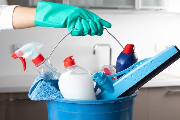 Weserve, cleaning blue bucket with cleaning products inside it held by a person wearing a blue glove with a white wall background and brown floor